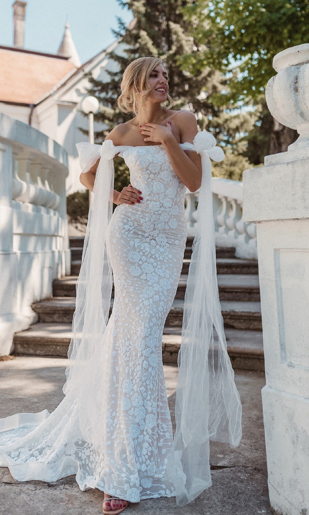 A woman in a strapless Tina Holly BB010 white wedding gown with lace and sheer train stands outdoors on stone steps by white railings and lush greenery.